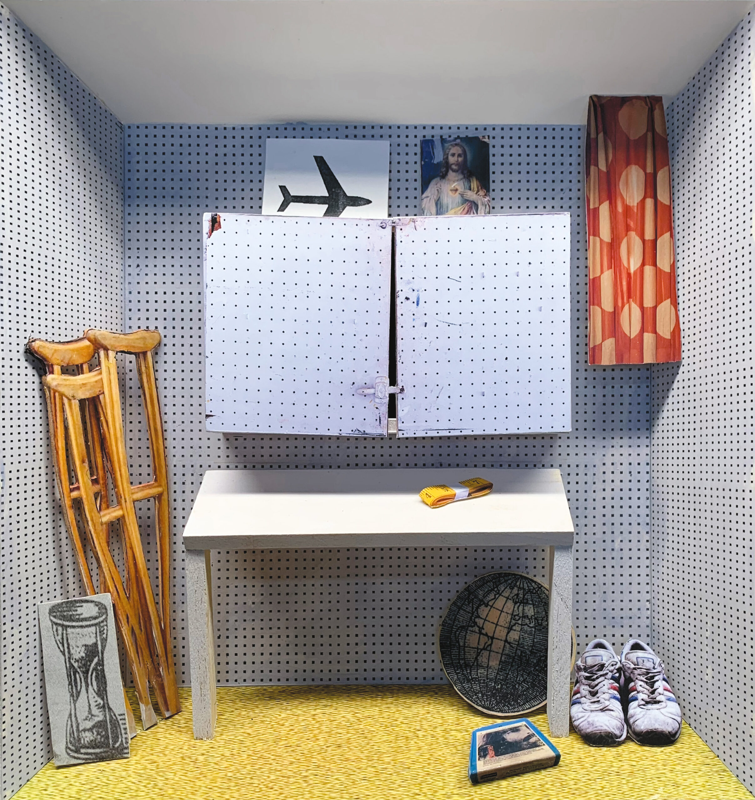 Les murs d'une petite pièce sont recouverts de panneaux perforés blancs. Le plancher de la pièce a différentes teintes de jaune. Une table blanche est au centre de la pièce. Un ruban à mesurer jaune est sur son dessus. En haut de la table, une armoire en panneaux perforés blancs est fixée au mur. Un dessin d'un avion et une illustration de Jésus sont en haut de l’armoire. À gauche de la table, trois photographies tailles réelles de béquilles en bois et d'un sablier sont appuyées contre le mur. À droite de la table, il y a une photographie taille réelle d'une paire de chaussures, une boite bleue et une image du globe de la Terre.