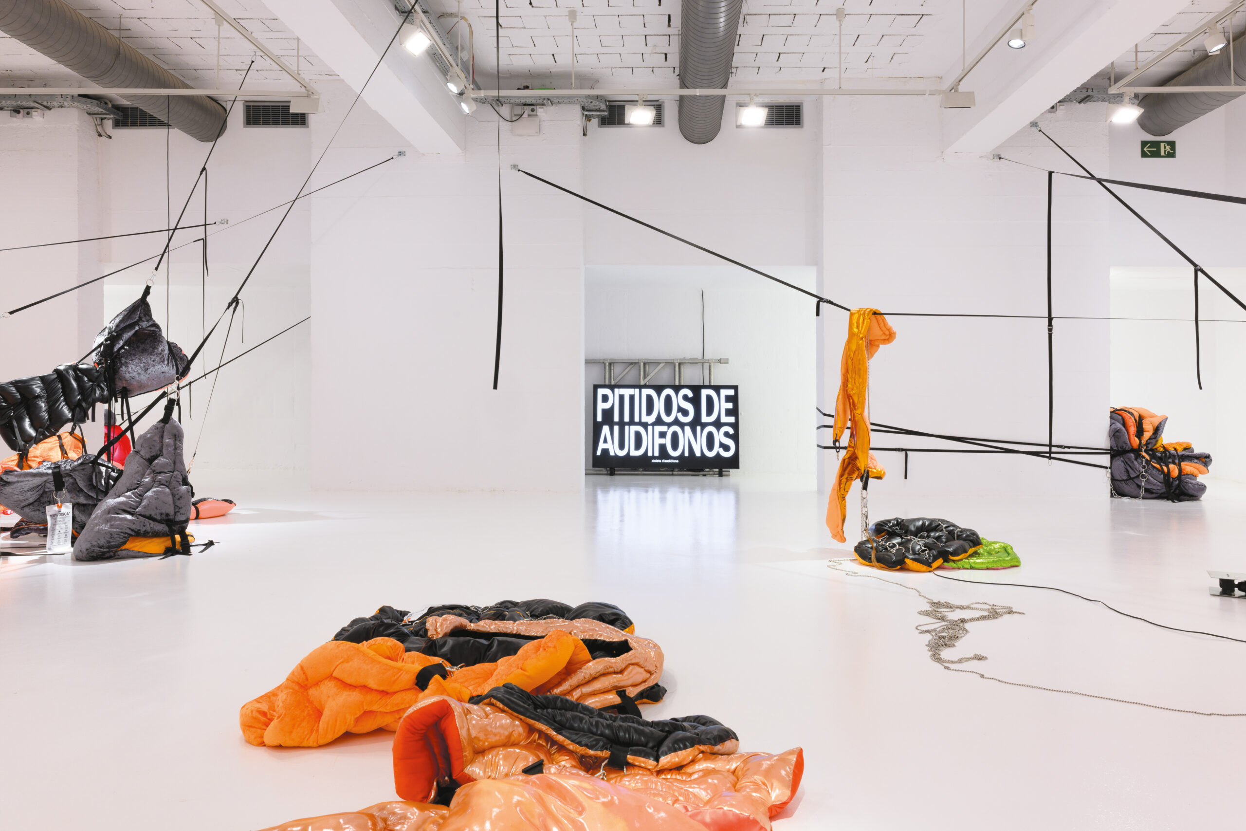 Des tissus matelassés roses, oranges, jaunes, verts et noirs sont éparpillés dans une salle blanche. À droite et à gauche de la pièce les tissus sont suspendus dans les airs à l'aide de sangles. Au centre de la pièce, les tissus ont été placés au sol. Au fond de la pièce, un panneau noir affiche PITIDOS DE AUDIFONOS en lettres blanches.