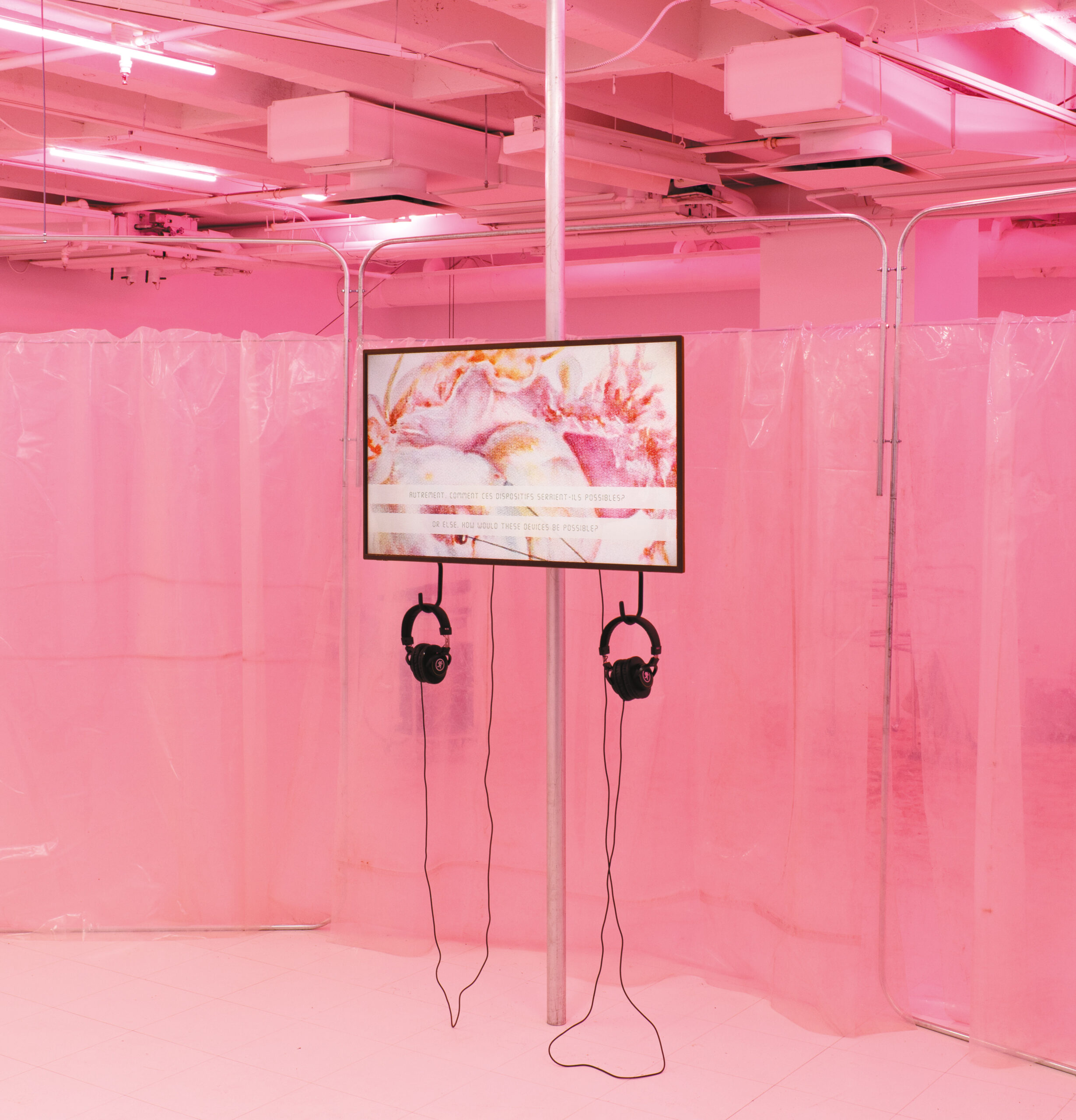 Dans une salle peinturée en rose, une télévision est fixée à un poteau de métal. La télévision projette des formes ondulées roses, rouges, blanches et orange. Deux casques d’écoute noirs sont branchés à la télévision.