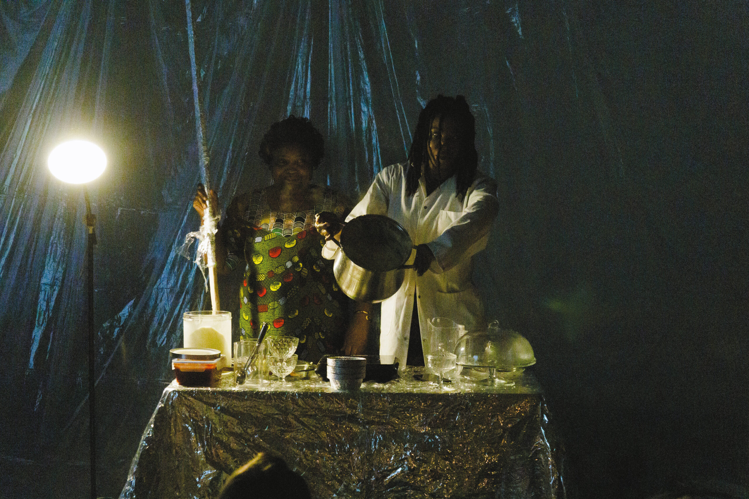 Dans une pièce sombre, deux personnes afrodescendantes sont derrière une table recouverte d'une nappe argentée. La personne de droite a des habits colorés et tient un long bâton dans la main gauche. La personne de gauche porte un sarrau blanc et montre le contenu d'un chaudron devant elle. La table comporte plusieurs gamelles. À droite de la table, un lampadaire émet de la lumière vers l'avant.